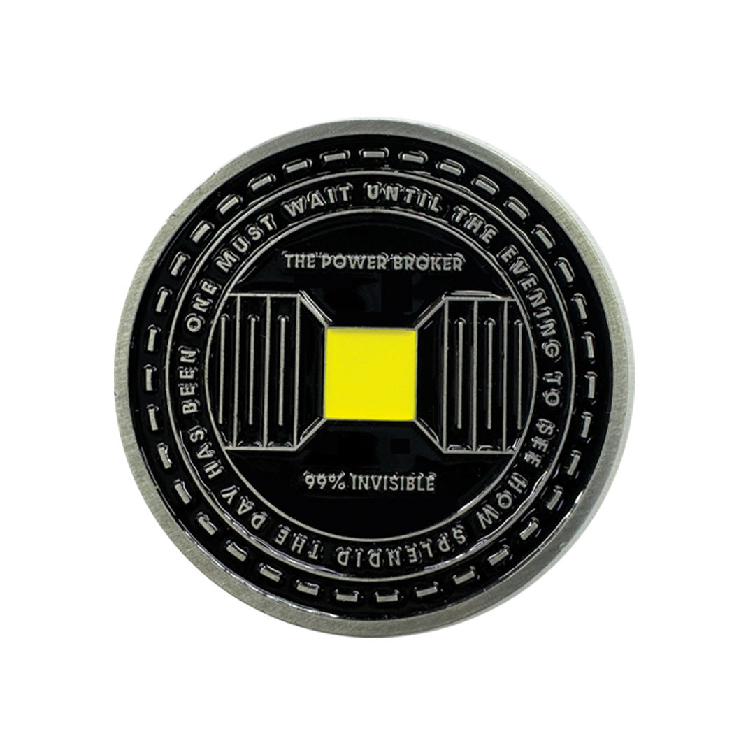 One side of black coin featuring a central yellow square and text that reads 'THE POWER BROKER,' '99% INVISIBLE,' and 'ONE MUST WAIT UNTIL THE EVENING TO SEE HOW SPLENDID THE DAY HAS BEEN'