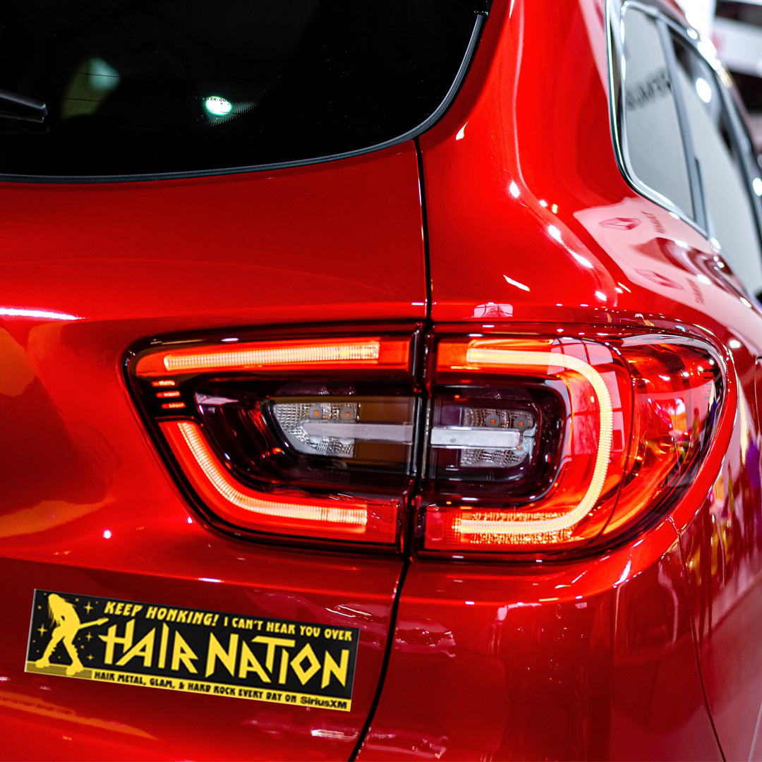 Close-up of the rear light and bumper of a red car, featuring a humorous 'Hair Nation' bumper sticker related to rock music.