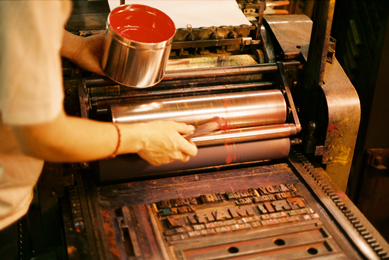 A person applying red ink to a poster printing press roller, preparing for printing on a vintage machine.