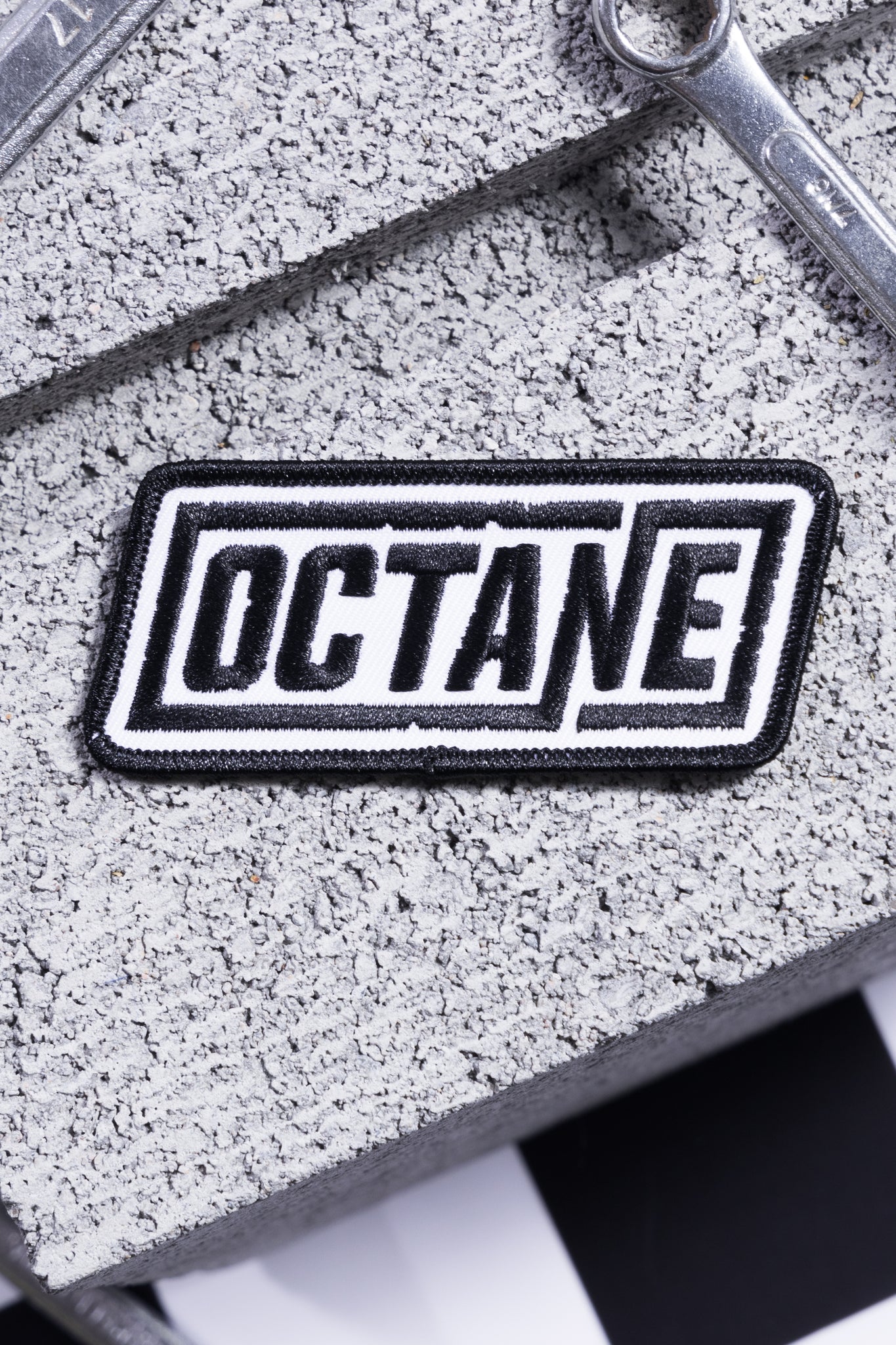 Black and white embroidered patch with the word 'OCTANE' on a textured gray surface.