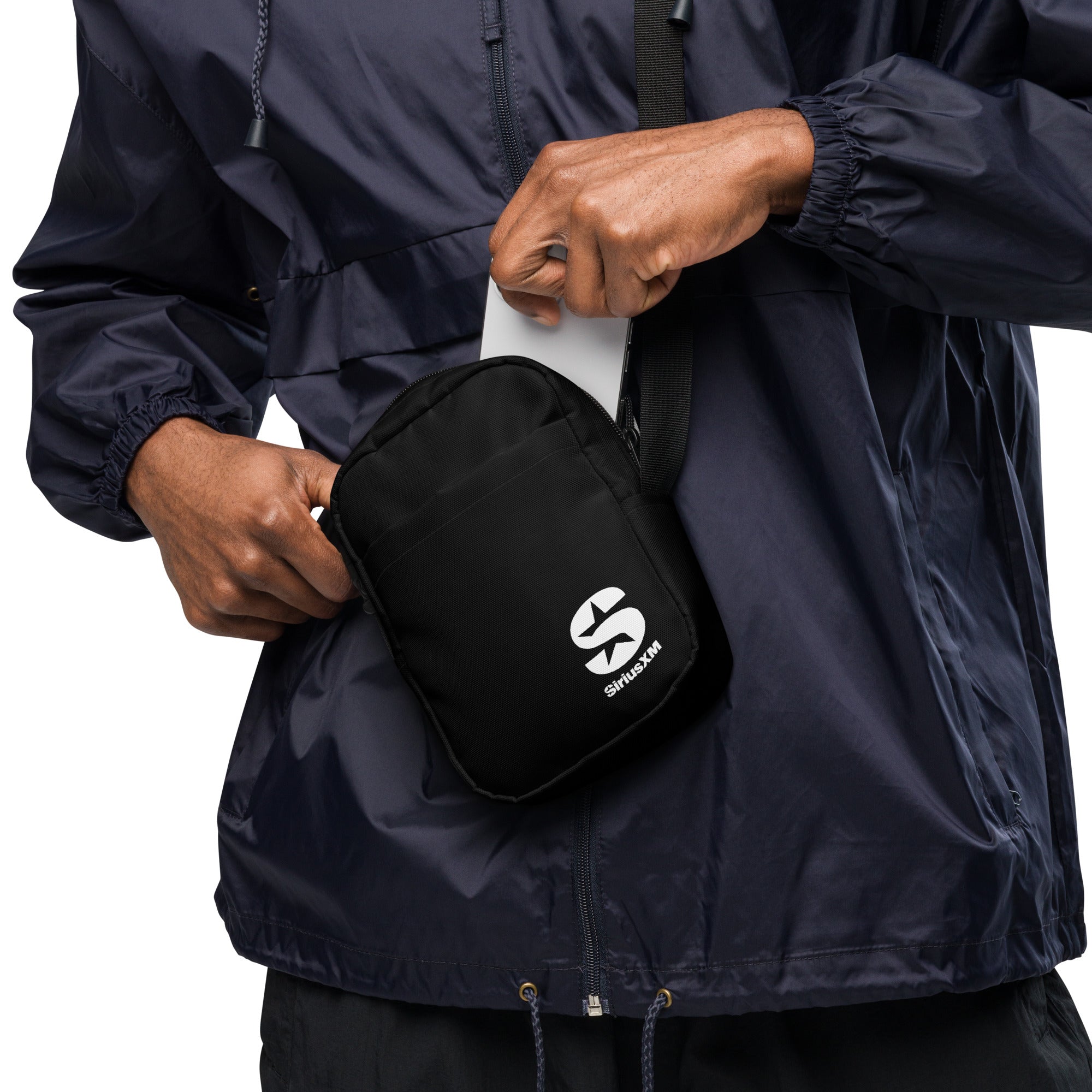 The midsection of a person wearing a navy windbreaker, placing a smartphone into a black 'SiriusXM' crossbody utility bag. The compact bag features the SiriusXM "S" logo in white on the front and has an adjustable strap for convenience.