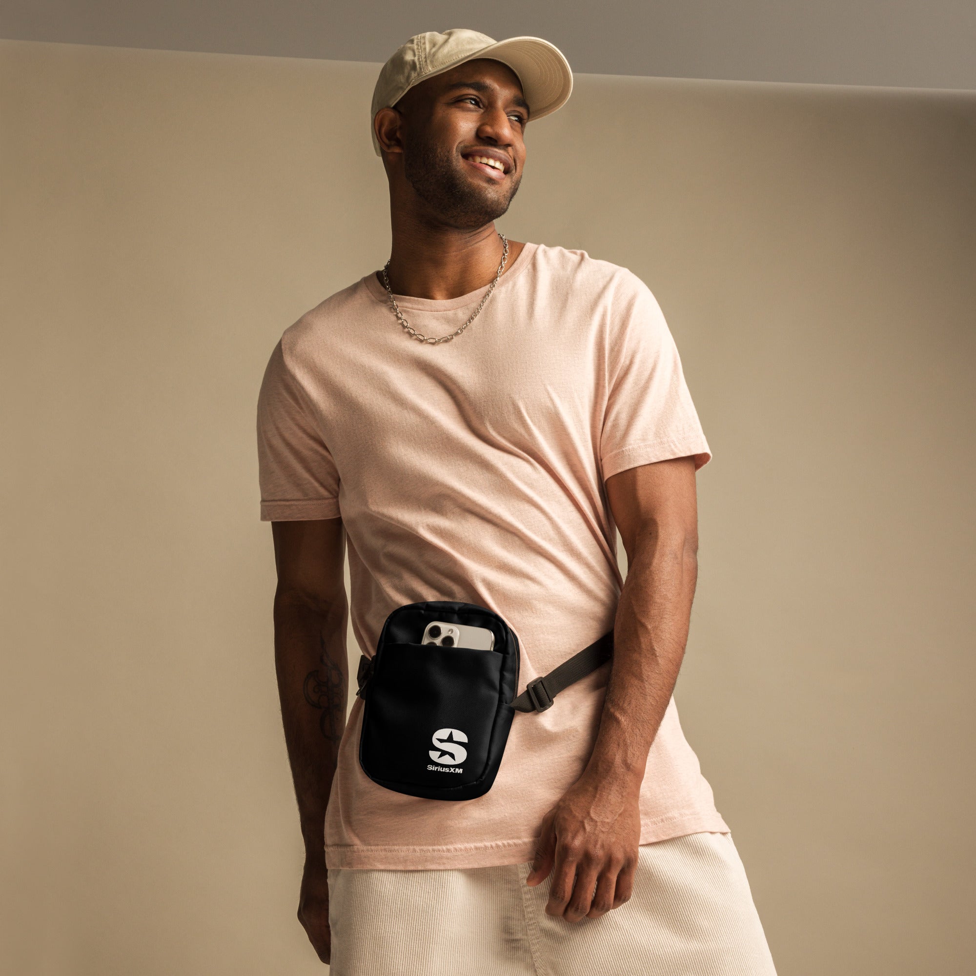 A man in a casual beige outfit wearing the 'SiriusXM' utility bag as a waist or fanny pack. The bag is secured around his waist with an adjustable strap and holds a smartphone in the front pocket. He is smiling and looking off to the side.