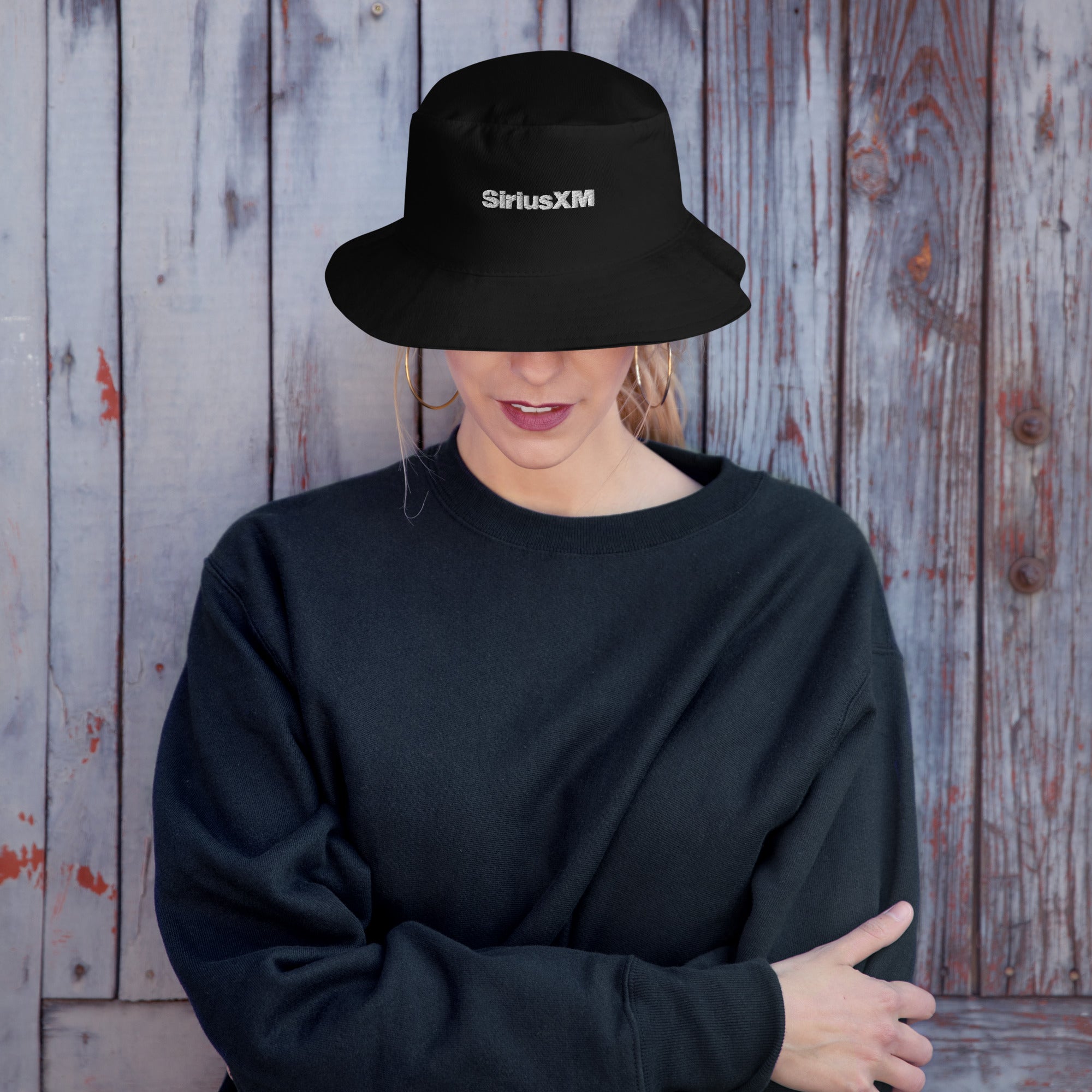 A woman standing against a rustic wooden background wearing a black bucket hat with a structured, wide-brim design featuring the 'SiriusXM' logo embroidered in white on the front. The hat sits low on the wearer’s head, partially obscuring her face.