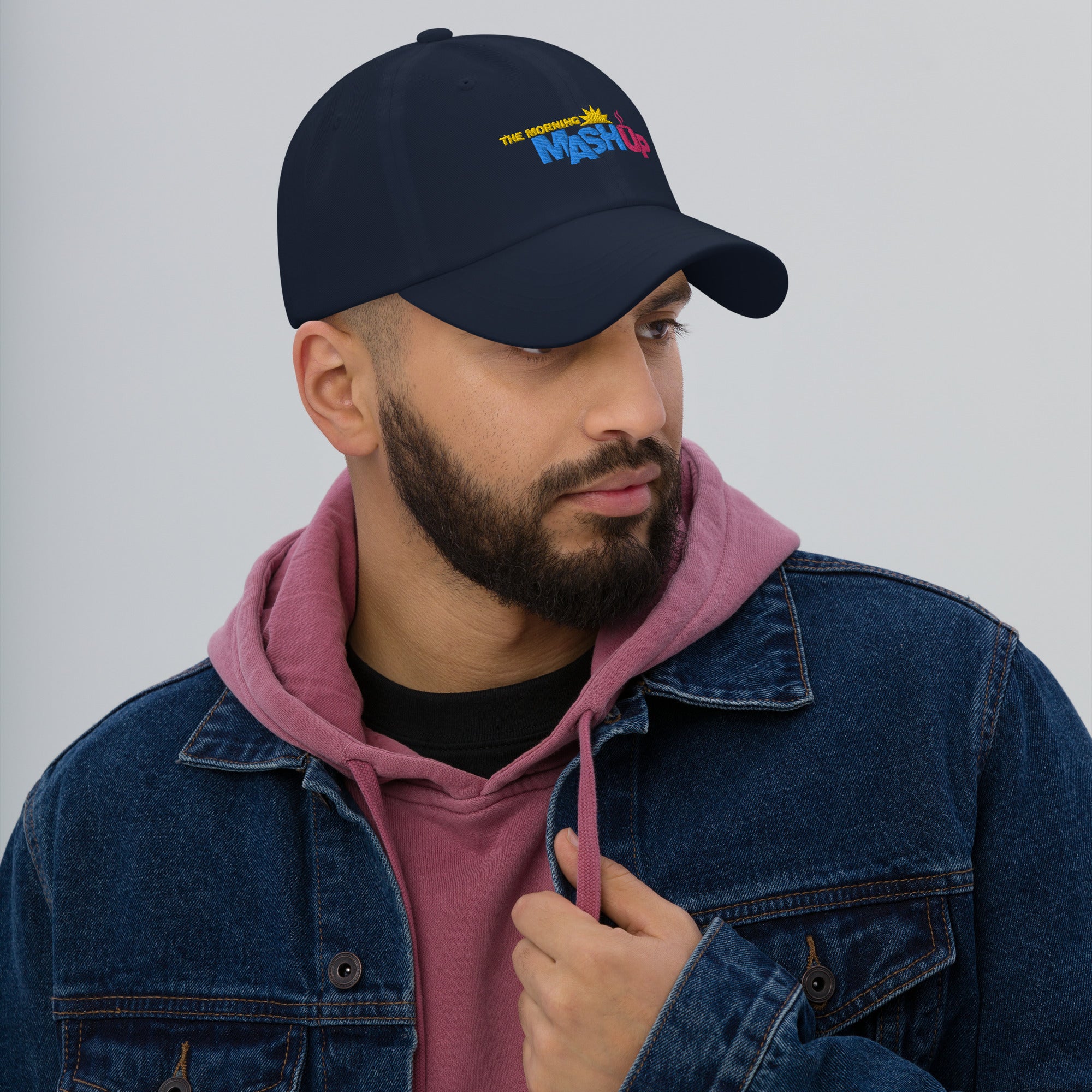 A man wearing a dark baseball cap with a colorful logo, a pink hoodie, and a denim jacket.