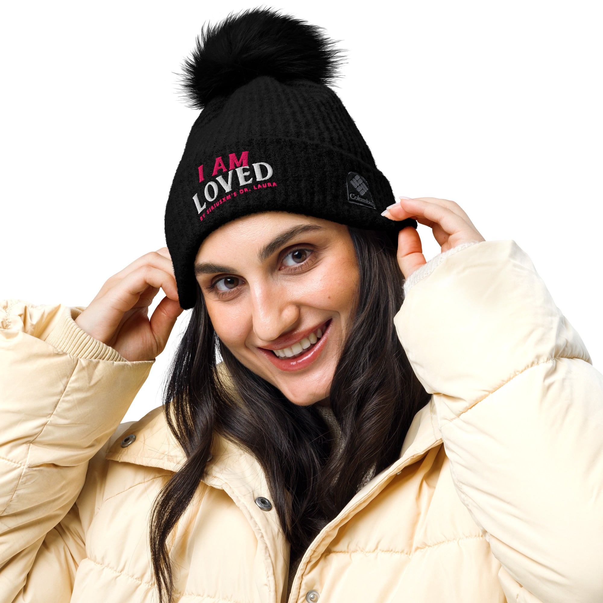 Smiling woman with dark hair wearing black knit beanie with a pom-pom on top. Hat features embroidered message that says 'I AM LOVED,' with 'I AM' in pink and 'LOVED' in white lettering. Below, it says 'BY SIRIUSXM'S DR. LAURA' in smaller pink text. Model is holding beanie's cuff, as if she just put it on. 