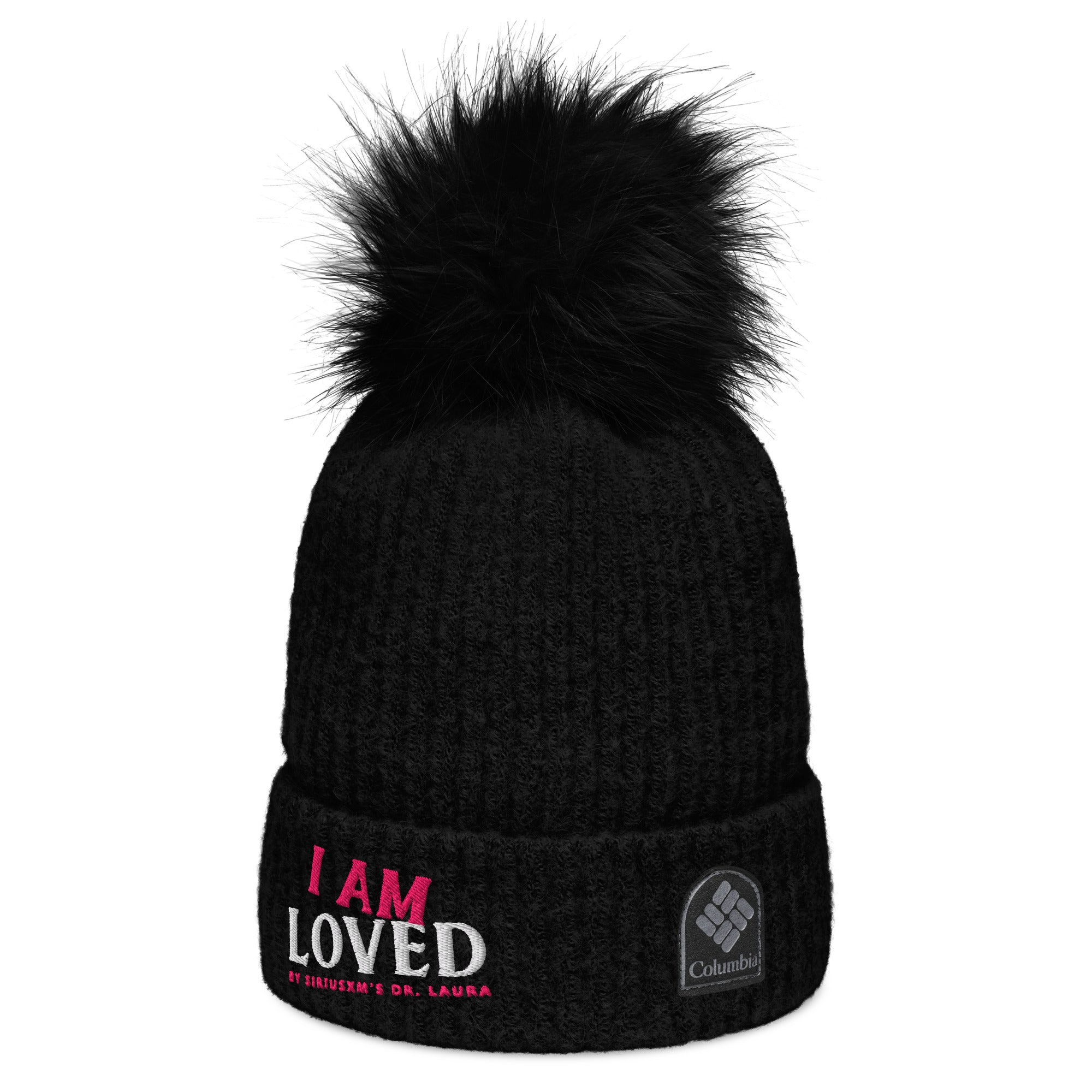 Side view of a black, ribbed beanie hat with matching, fluffy pom-pom on top and embroidered text on the cuff reading 'I AM LOVED,' with 'I AM' in pink and 'LOVED' in white lettering. Below, it says 'BY SIRIUSXM'S DR. LAURA' in smaller pink text. The beanie also features a 'Columbia' logo patch on the cuff.