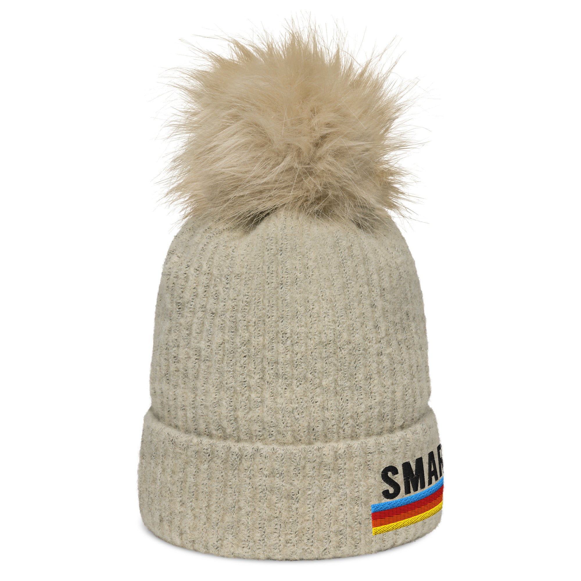 A cream-colored knit pom-pom beanie with a fluffy tan pom-pom on top. The front side of the beanie shows part of the word 'SMARTLESS' with a rainbow stripe design, along with a folded cuff for added style.