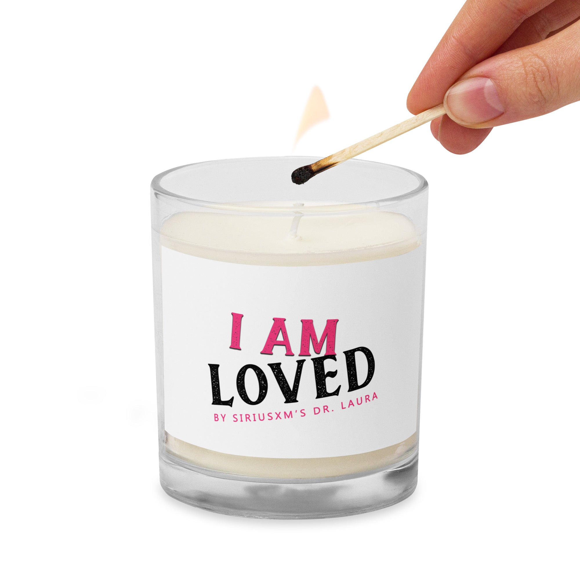 A clear glass jar candle with white soy wax inside. The label on the front reads 'I AM LOVED' in bold text, with 'I AM' in pink and 'LOVED' in black. Below, it says 'BY SIRIUSXM'S DR. LAURA' in smaller pink text. There is a partially visible hand lighting the candle with a matchstick in upper right corner.  