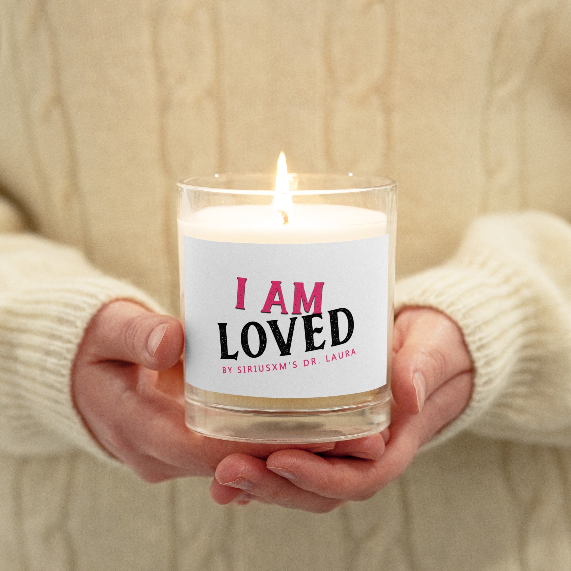 A lit clear glass jar candle with white soy wax inside. The label on the front reads 'I AM LOVED' in bold text, with 'I AM' in pink and 'LOVED' in black. Below, it says 'BY SIRIUSXM'S DR. LAURA' in smaller pink text. The candle being held in the hands of someone wearing a white knit sweater. 