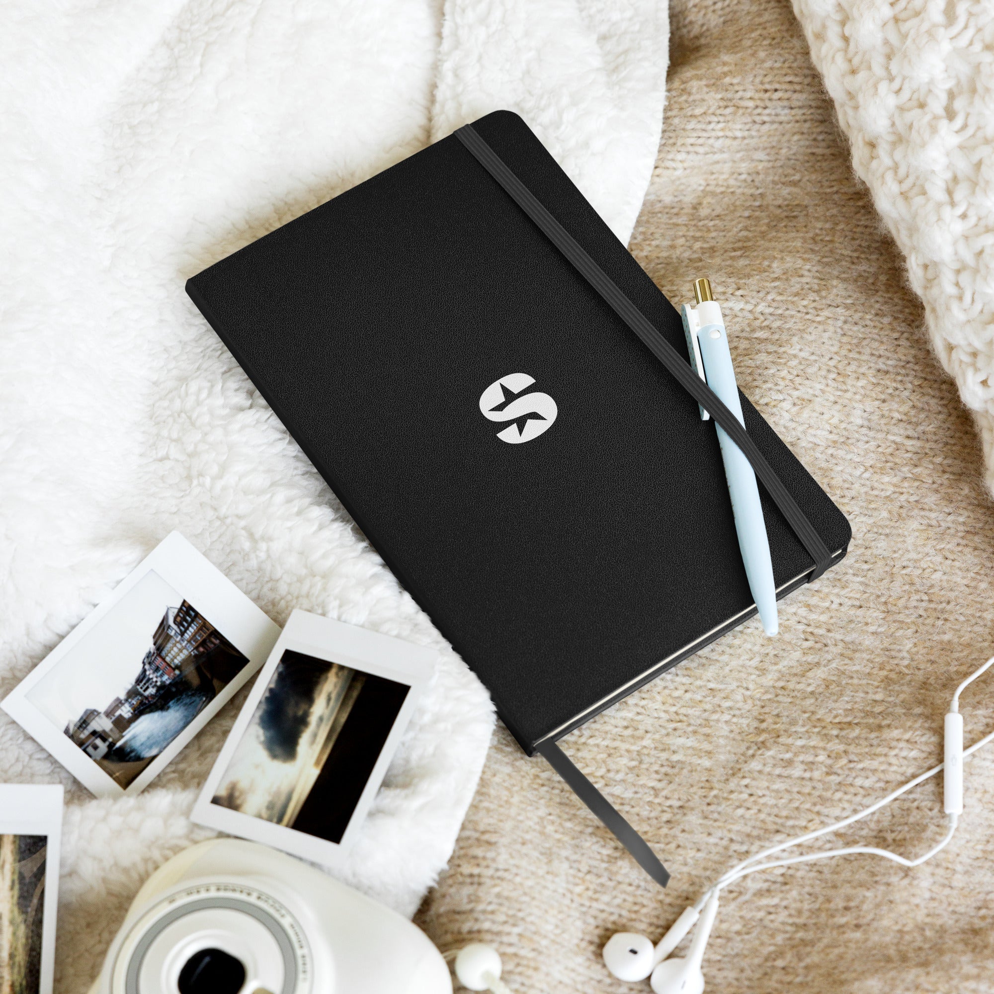 The same black 'SiriusXM' notebook resting on a soft, cozy surface alongside a light blue pen, instant film photographs, and a pair of white earphones, creating a relaxed and creative atmosphere.