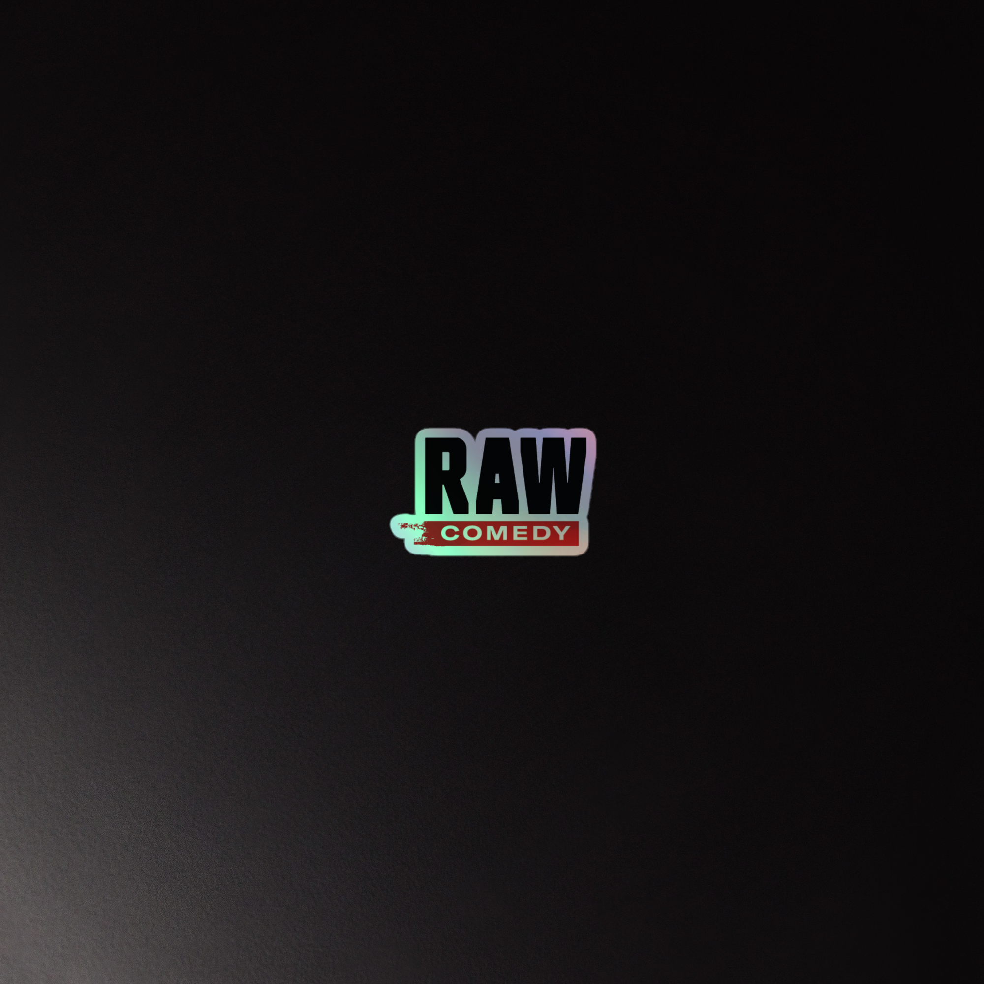 Holographic sticker of black and red 'RAW COMEDY' logo on a black surface.