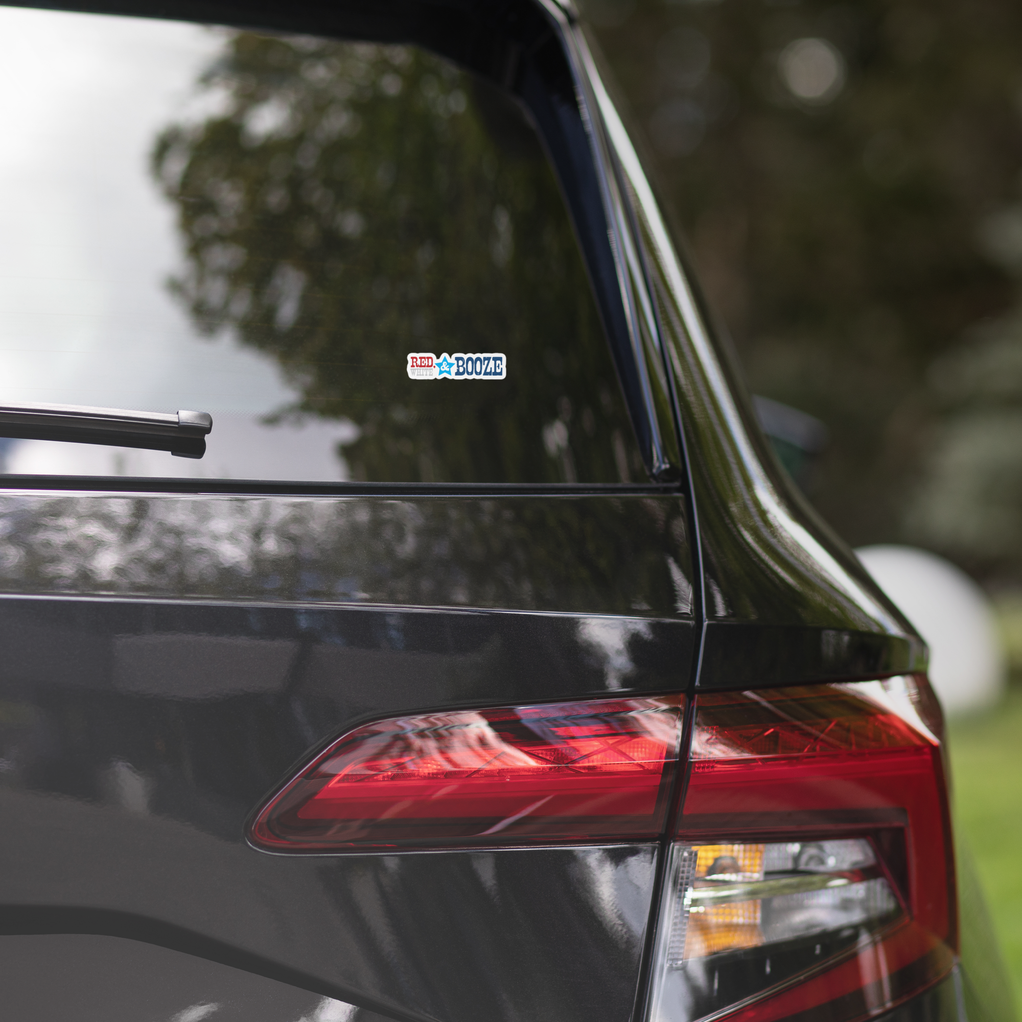 Close-up of a black car's rear window displaying a colorful 'RED, WHITE & BOOZE' sticker.