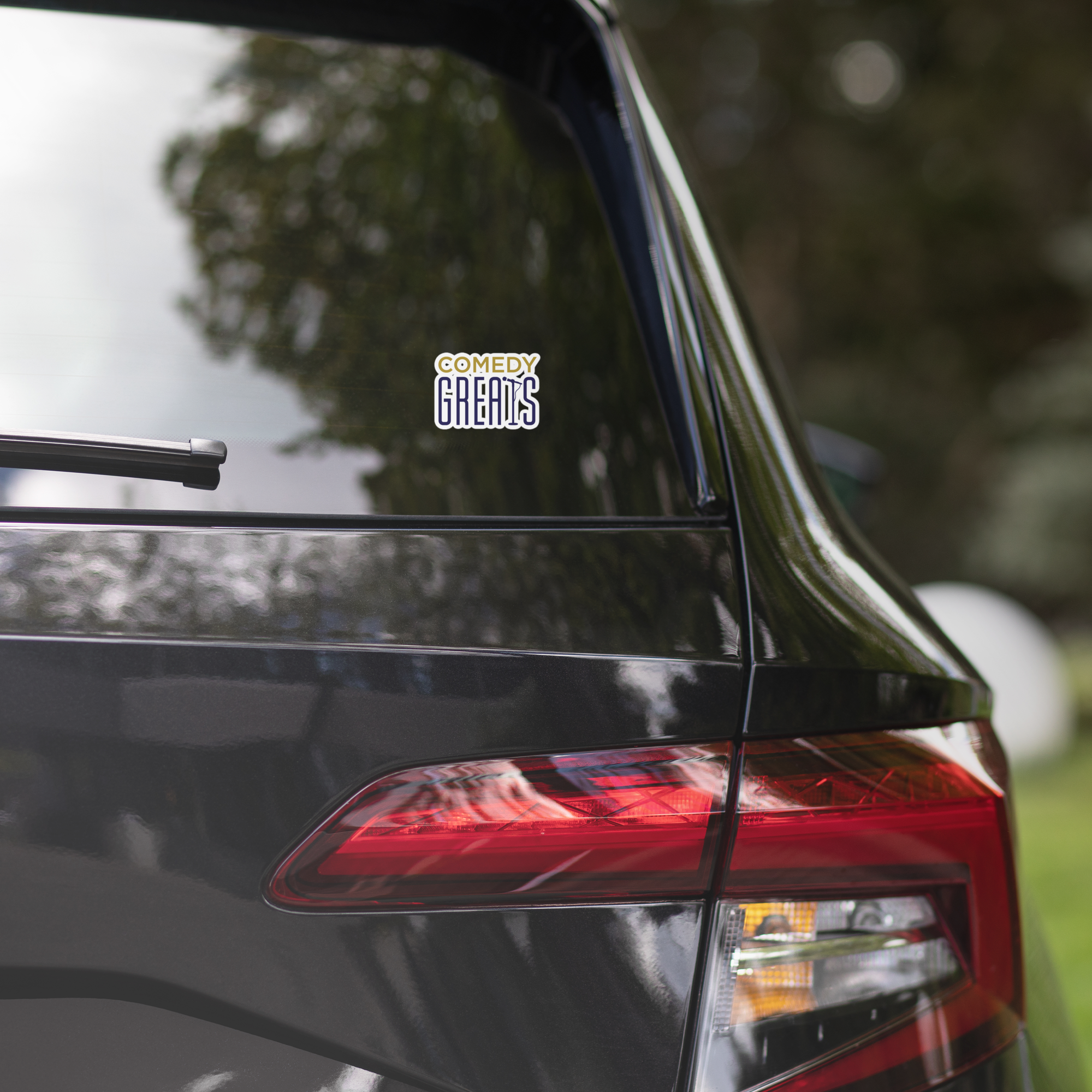 Close-up of a black car's rear window displaying a 'COMEDY GREATS' sticker.