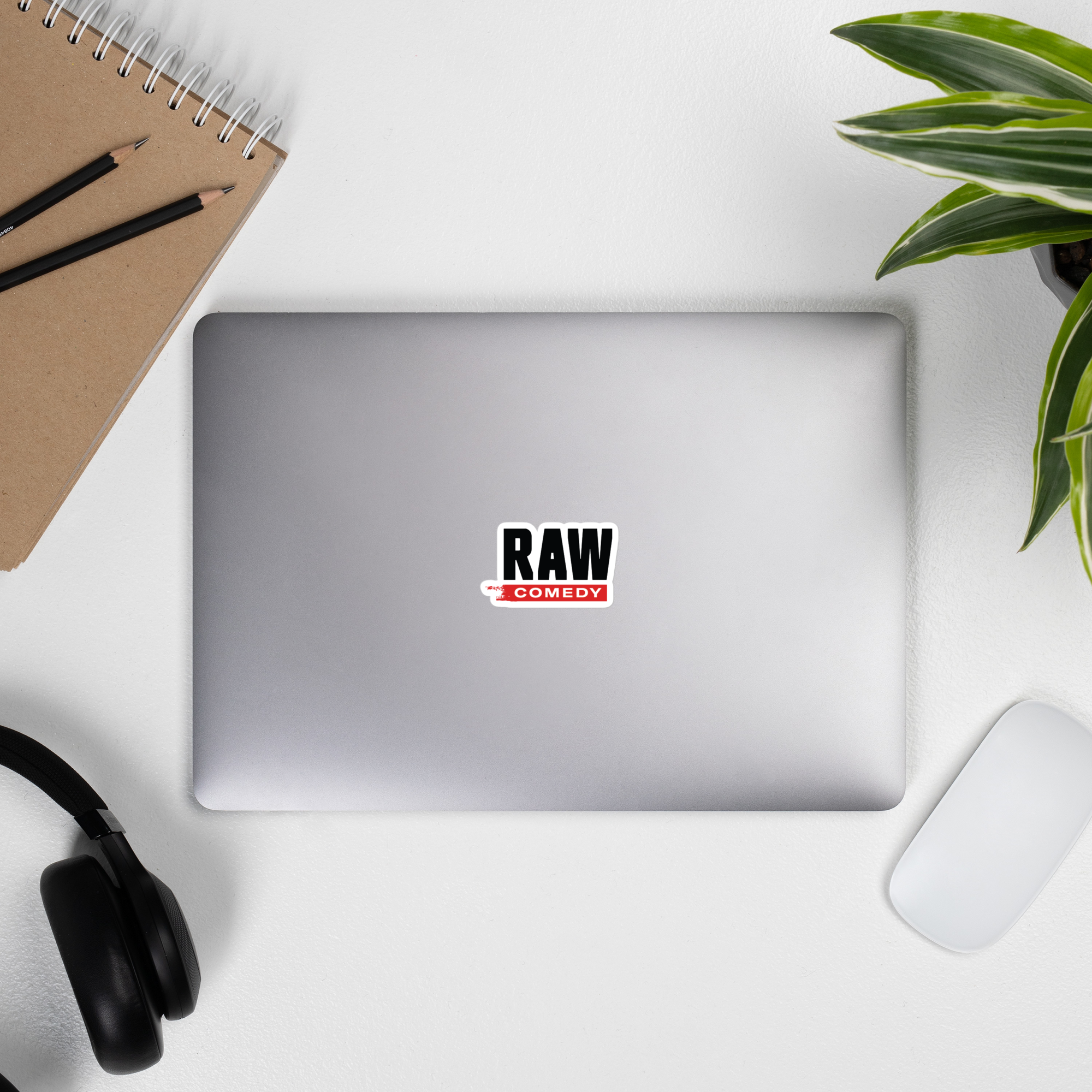 Flat lay of a laptop with a 'RAW COMEDY' sticker, notebook, pencils, headphones, and a plant on a white surface.