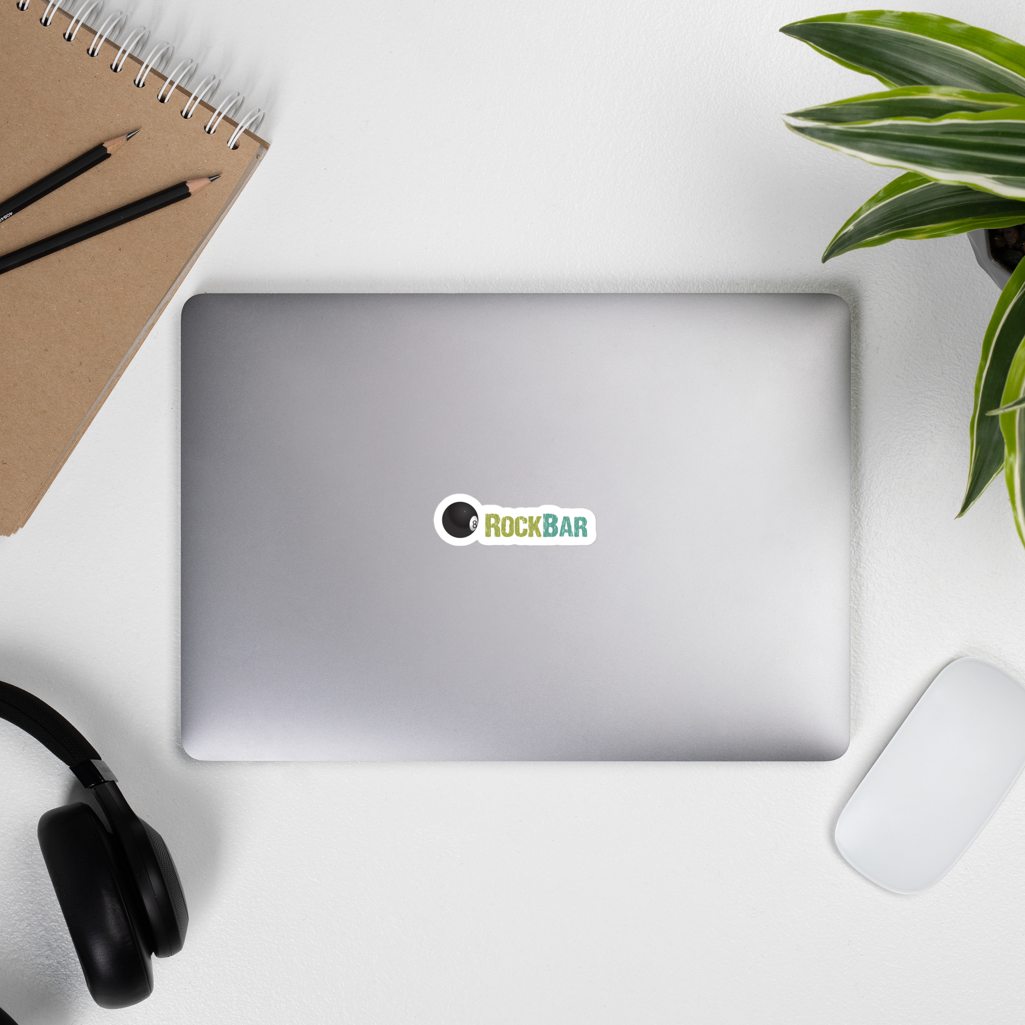 A laptop with a 'ROCKBAR' sticker, surrounded by a notebook, pencils, headphones, a plant, and a mouse on a white surface.