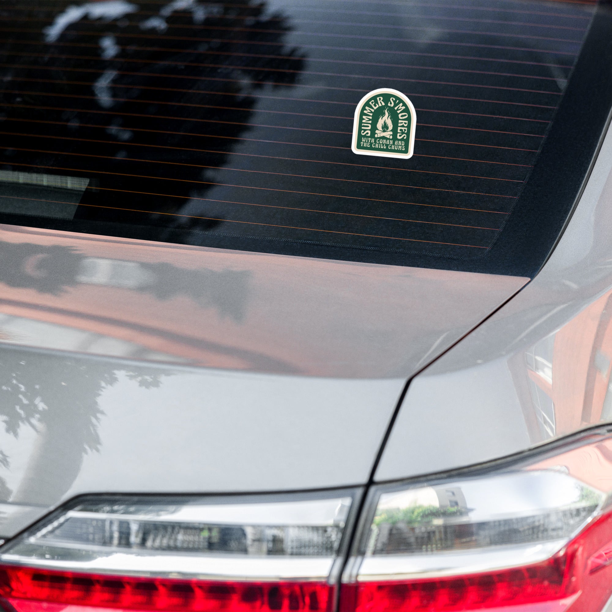 A close-up of a car's rear window displaying a sticker that reads 'SUMMER SWEEZE' with a graphic design, capturing reflections of the surroundings.