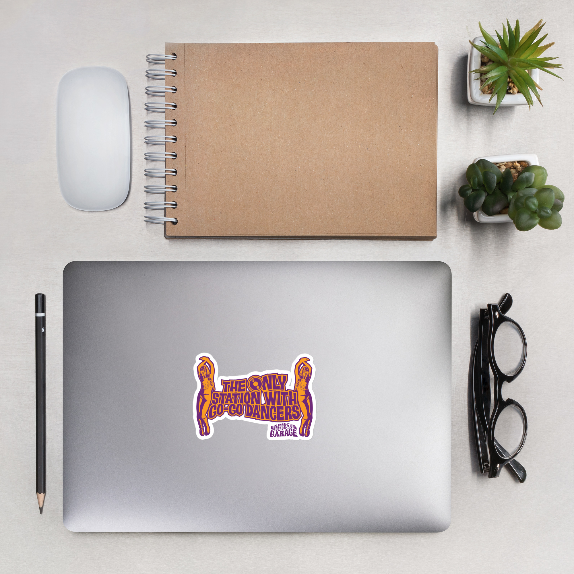 Flat lay of a workspace featuring a laptop with a colorful sticker, a pencil, a mouse, a notebook, and small potted plants.