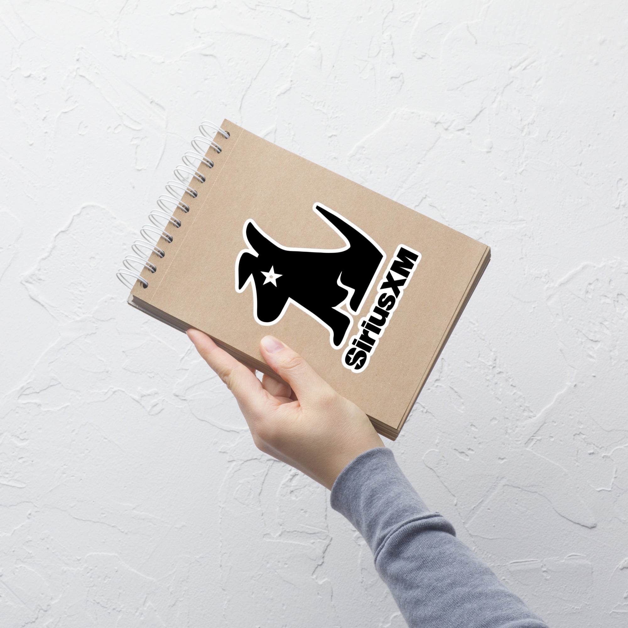 Hand holding a spiral-bound notebook displaying black Stella 'SiriusXM' sticker on it.