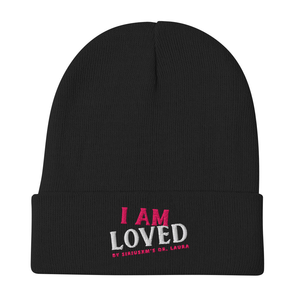 A black, cuffed knit beanie laying flat on a white surface featuring embroidered text that reads 'I AM LOVED,' with 'I AM' in pink and 'LOVED' in white lettering. Below, it says 'BY SIRIUSXM'S DR. LAURA' in smaller pink text.