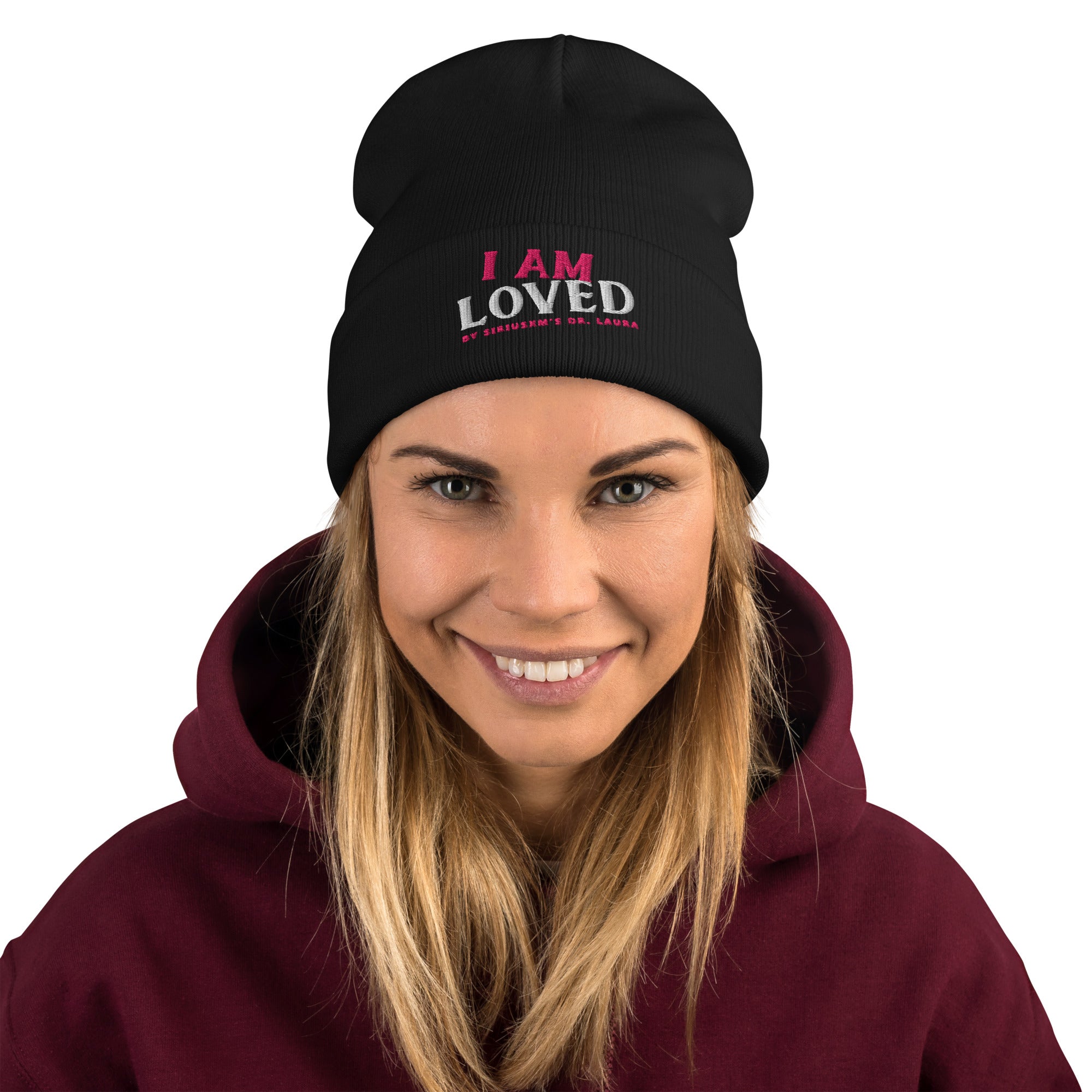 A blond haired, smiling woman wearing black knit beanie with an embroidered text that says 'I AM LOVED,' with 'I AM' in pink and 'LOVED' in white lettering. Below, it says 'BY SIRIUSXM'S DR. LAURA' in smaller pink text.