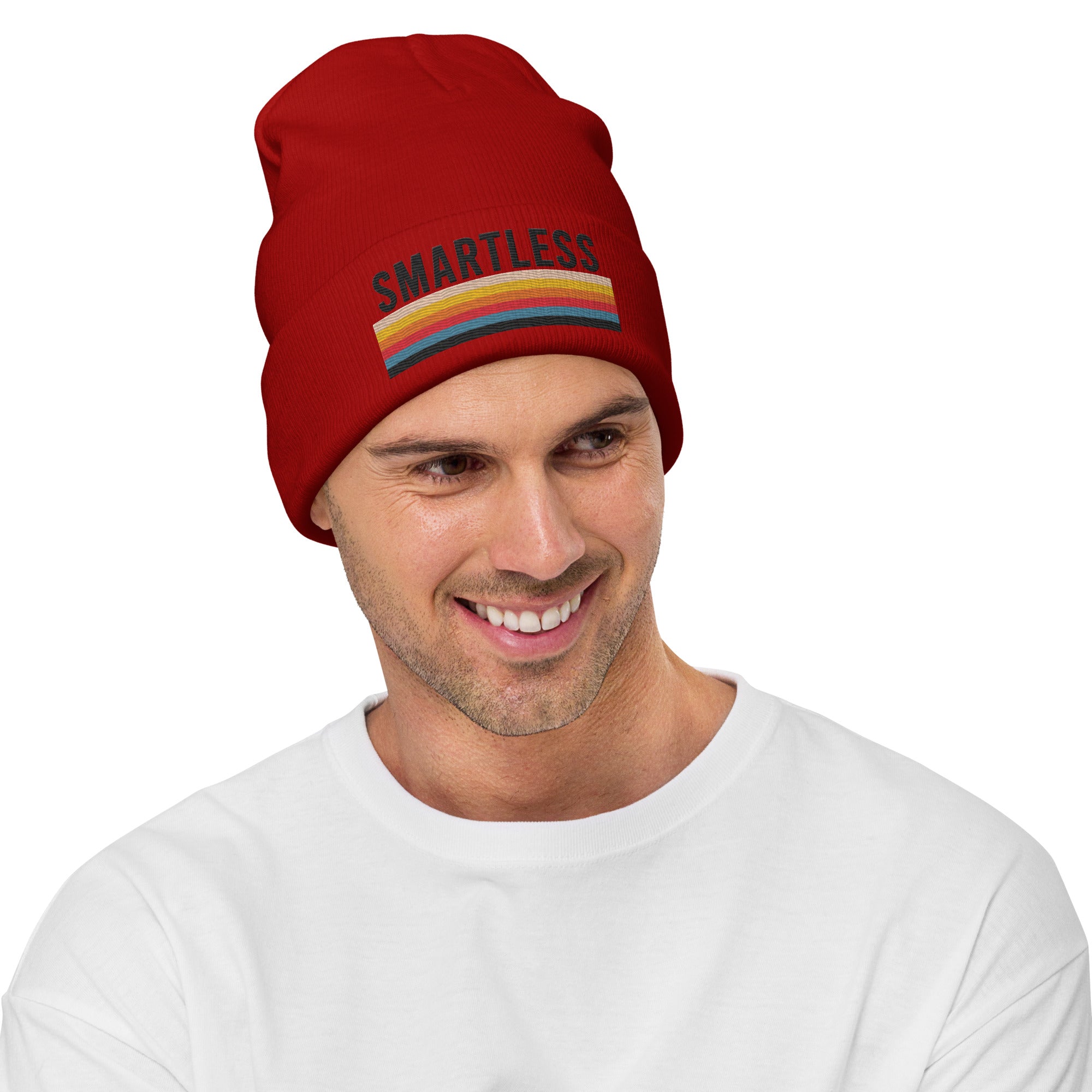 A smiling man wearing a red knit beanie featuring the word 'SMARTLESS' in black capital letters above a horizontal multicolored stripe pattern. He is against a plain white background.