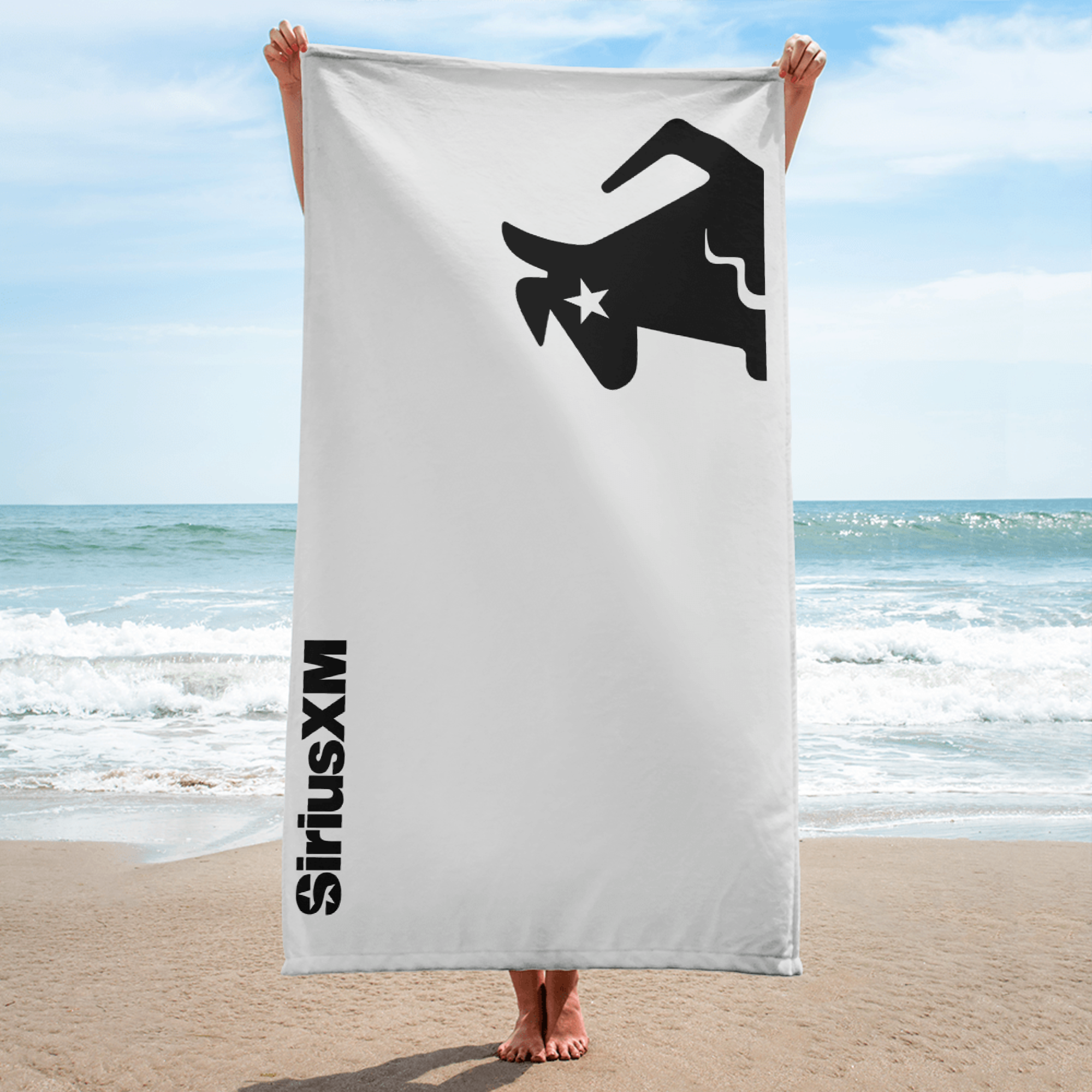 A person holding a large white beach towel with a black Stella deisgn and the text 'SiriusXM' in black on the side,  model standing on the beach.