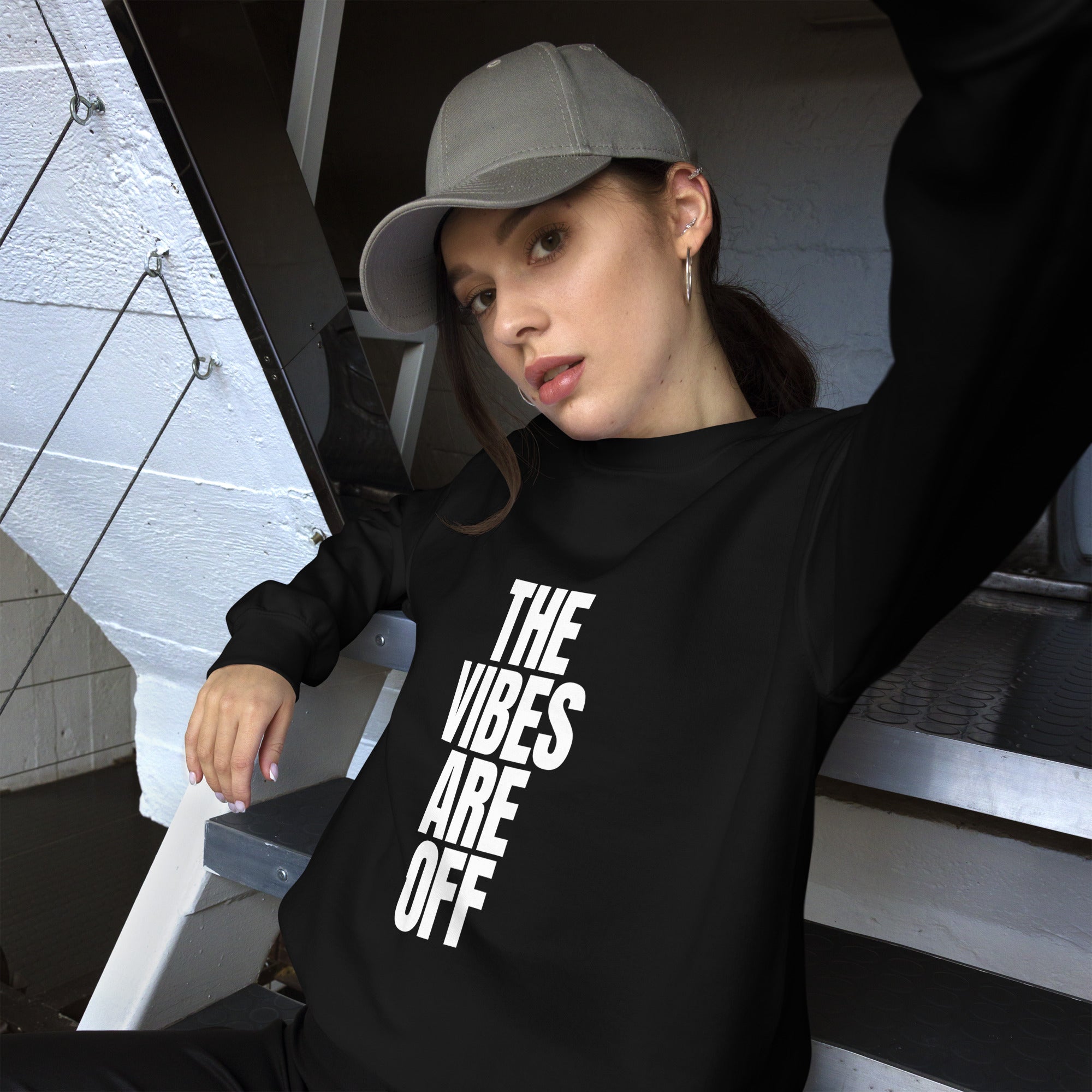 A woman reclining on stairs, wearing a grey baseball hat and black crewneck sweatshirt featuring bold white text that reads 'THE VIBES ARE OFF' in a stacked design.