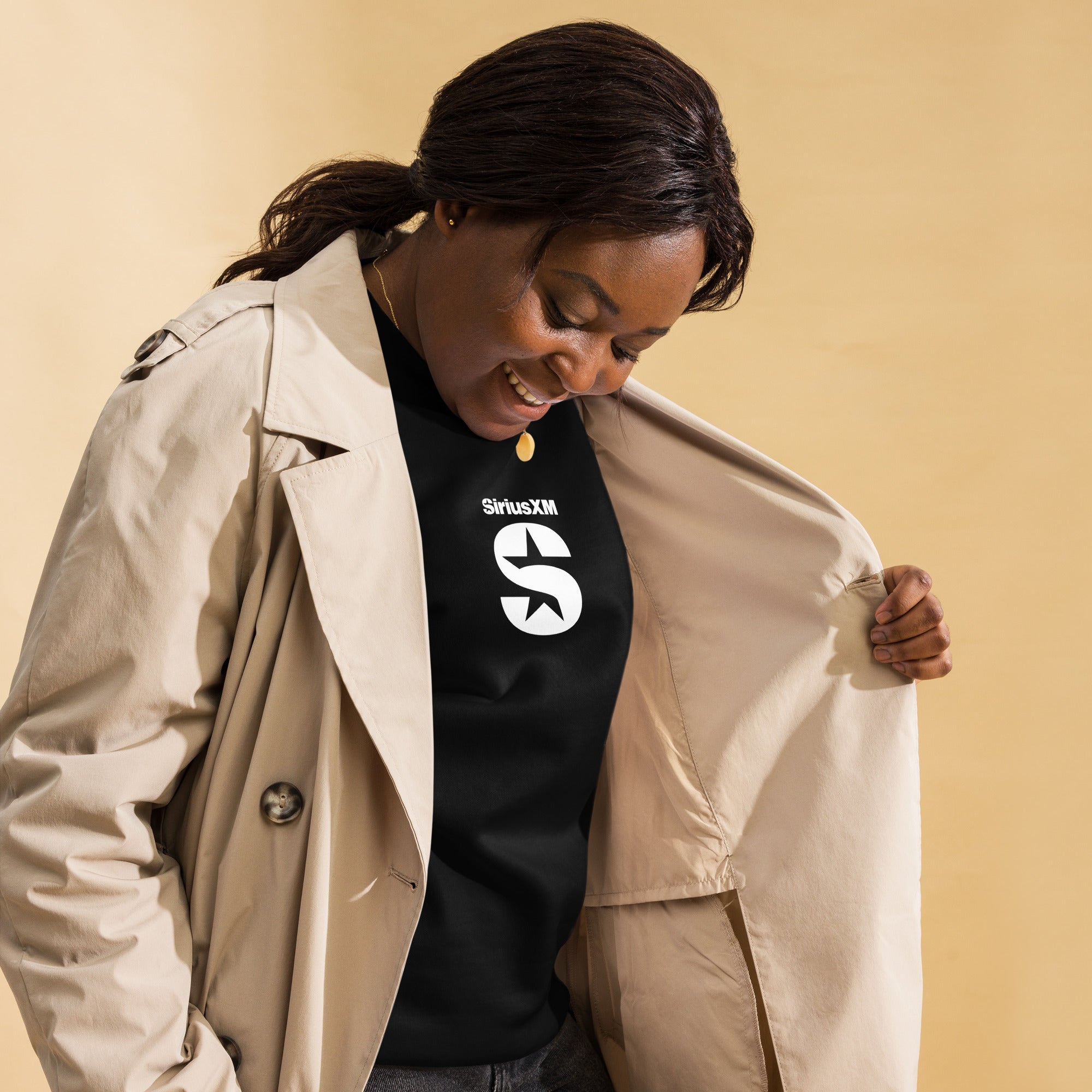 A woman wearing a black 'SiriusXM' sweatshirt layered under a beige trench coat. She is smiling and opening her coat to reveal the sweatshirt’s white 'SiriusXM' logo, which features a bold 'S' with a star cutout. The background is a warm beige tone.