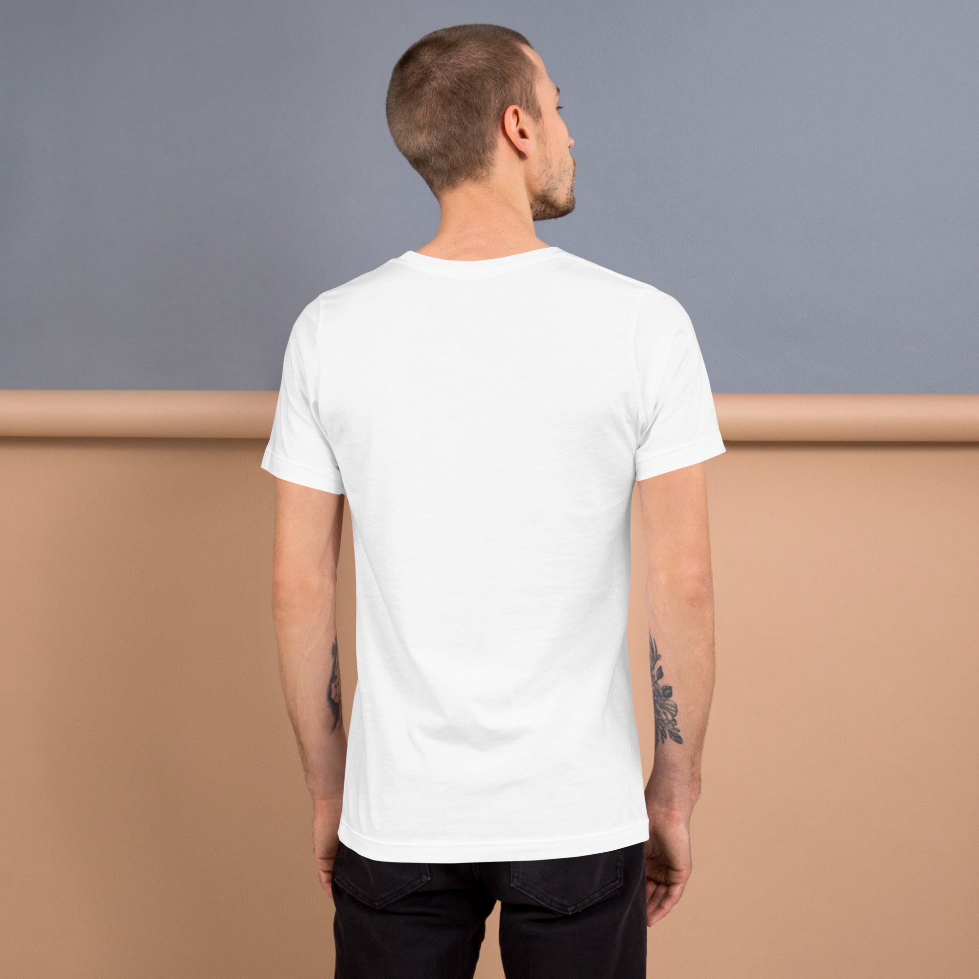 A man wearing a plain white t-shirt, viewed from the back. The shirt has a classic crew neck and short sleeves, with a smooth fit. He is also wearing black jeans, and his arms have visible tattoos. The background features a neutral-toned, two-tone setting