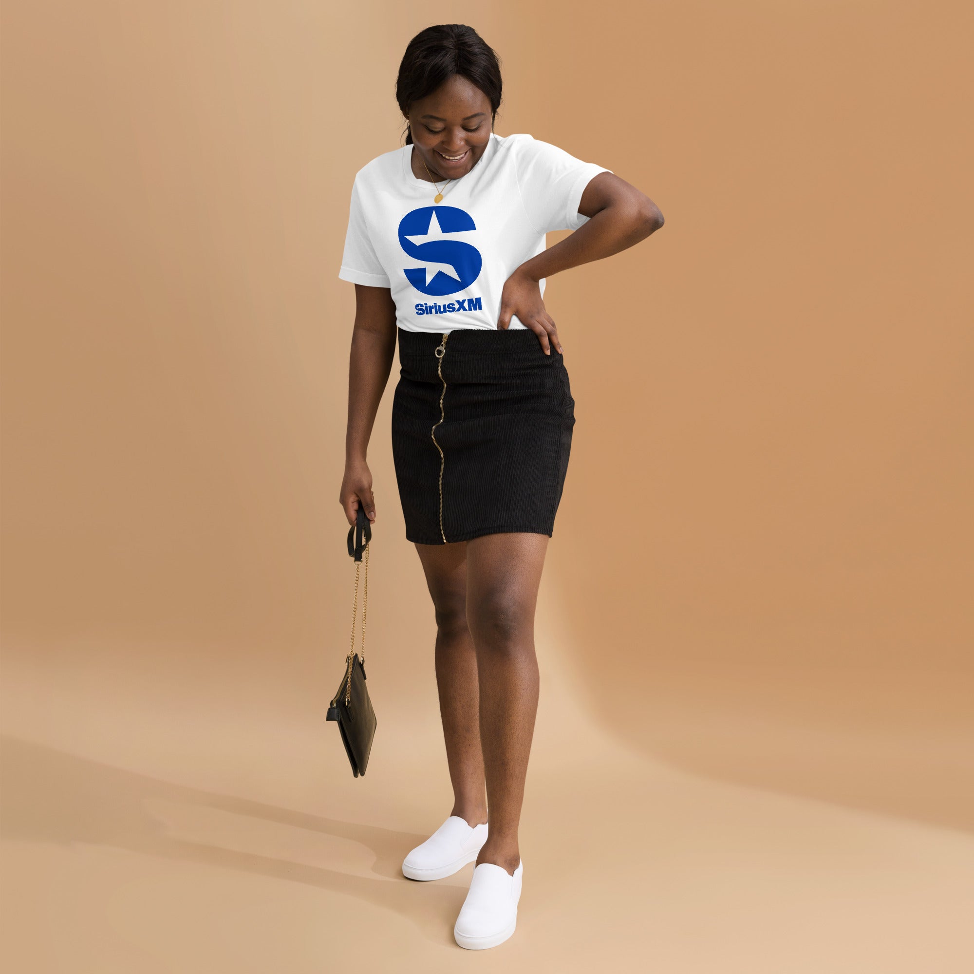 A woman wearing a white 'SiriusXM' t-shirt with a bold blue logo on the front. She is dressed in a black skirt with a gold zipper, white slip-on sneakers, and is carrying a black purse with a gold chain. She is smiling and adjusting her skirt, standing against a warm beige background.