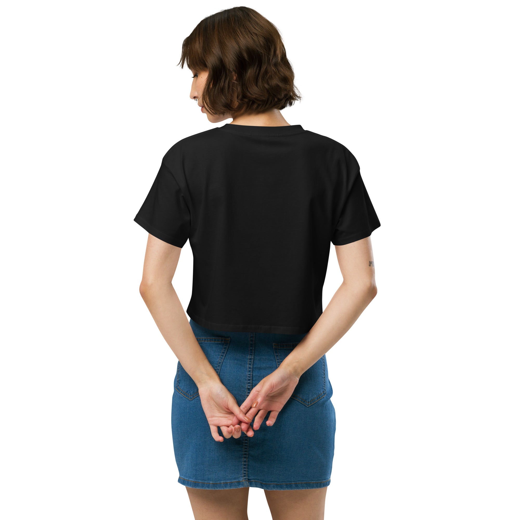 The back view of a woman wearing the black crop top. The design is plain on the back, and she is standing with her hands clasped behind her while wearing a blue denim skirt.