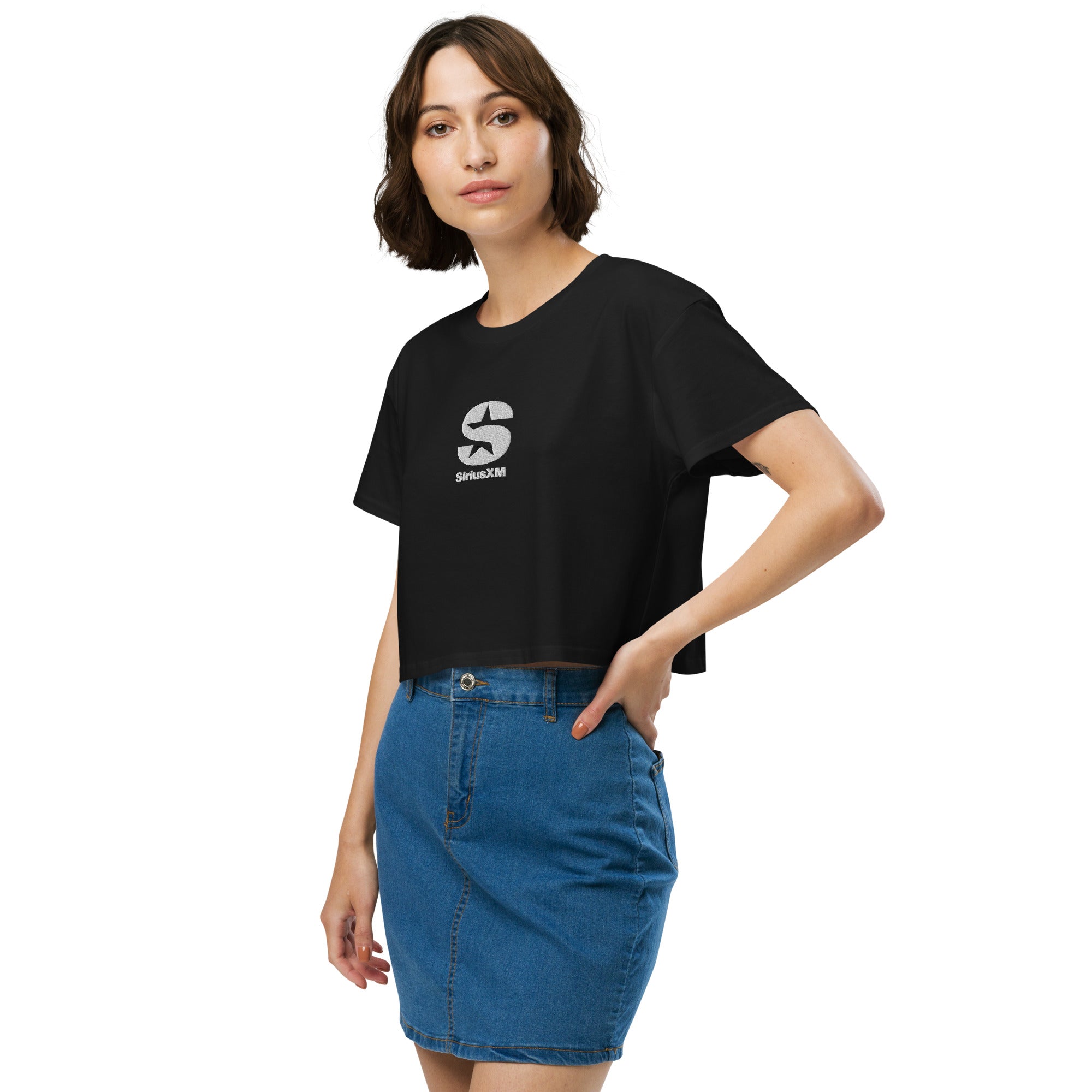 A woman wearing a black 'SiriusXM' crop top with a white embroidered logo on the chest. She pairs it with a blue denim skirt, standing with one hand on her hip.